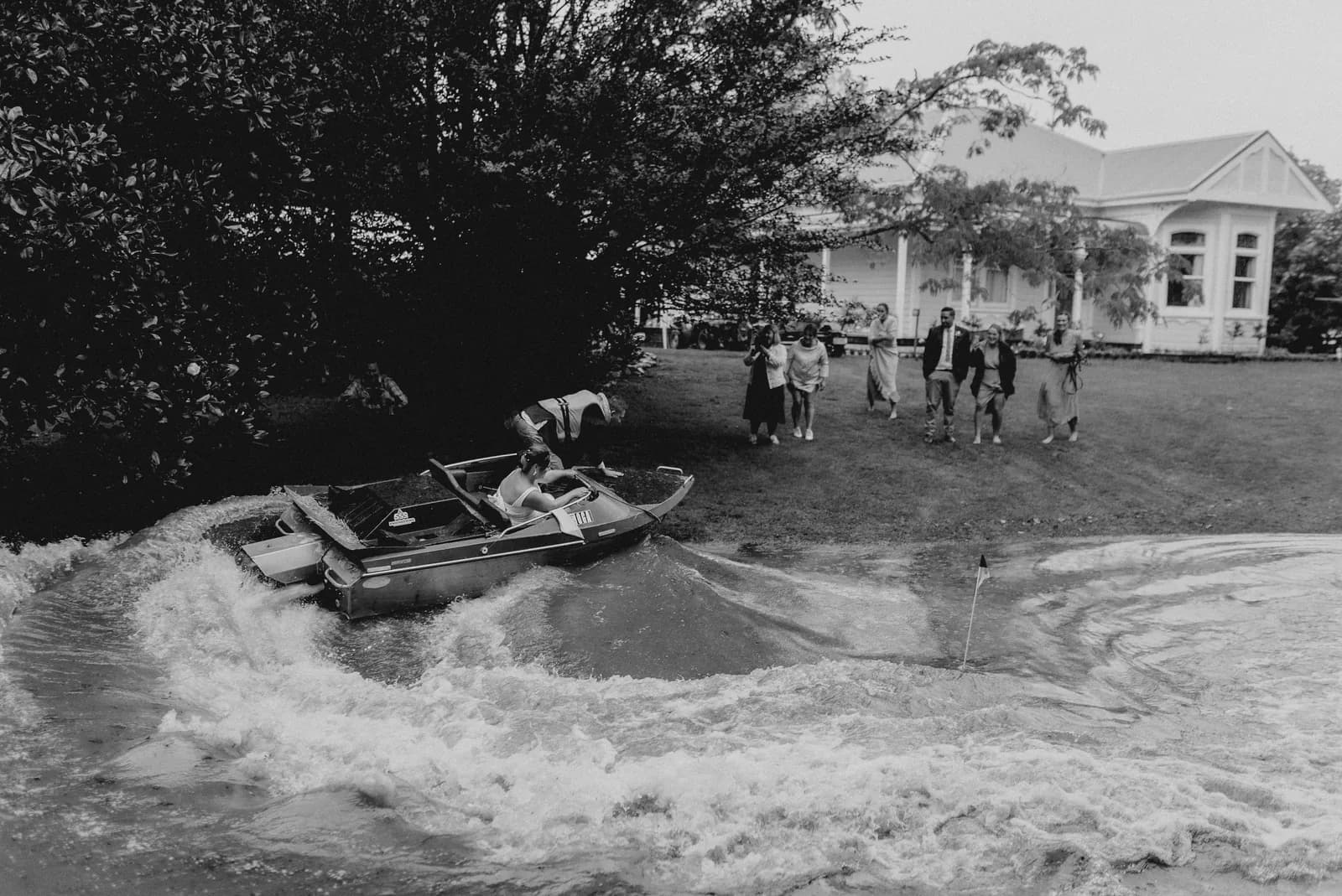 Black and white photo of a groom riding a jet ski through a flooded garden at his wedding